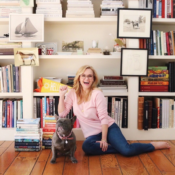 An image of Reese Witherspoon posing with a gray French bulldog in front of a bookshelf. 